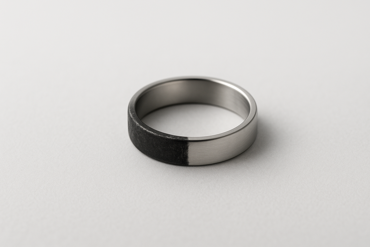 Pierścionek ze stali chirurgicznej leżący na jasnym tle, z wyraźnym kontrastem – połowa powierzchni srebrzysta i błyszcząca, druga połowa przyciemniona i matowa, symbolizująca możliwe czernienie metalu